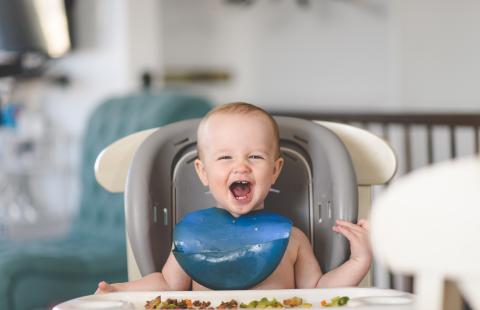 Infant boy in high chair wearing a bib and eating solid foods.