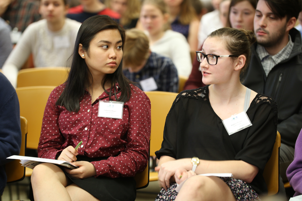 Two young women sit next to each other. One is looking ahead; the other is looking to her right at the other young woman.