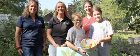 Three adults and two children in garden, holding produce and Plant a Row sign