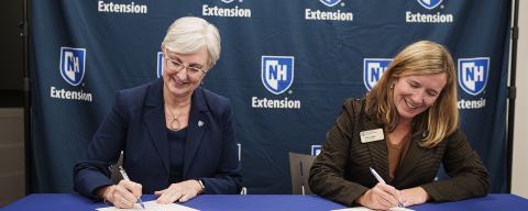 Two women signing papers at a table with blue UNH Extension logo banner in the background