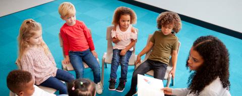 Young female school teacher reading a book to infant school children, sitting on chairs in a circle in the classroom listening.