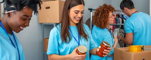 Cheerful young woman laughs along with her friend while volunteering in a community food bank. They are sorting through food donations.