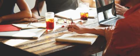 people at a table with papers, notebooks, an open laptop computer, and a beverage in a glass with ice 