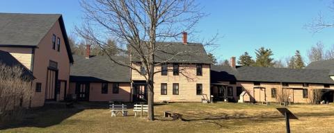 Several historic pinkish-gold farm buildings with black roofs stand around an open grassy field with leafless trees on a clear day