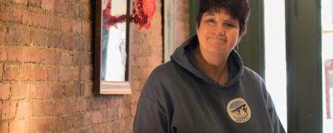 A woman in a grey hooded sweatshirt stands near a brick wall. She is facing the camera and smiling.