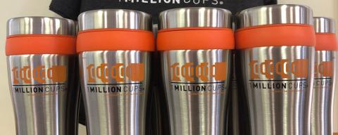 Five travel coffee mugs with the logo for 1 Million Cups in orange. Behind the mugs is a t-shirt with the logo for 1 Million Cups