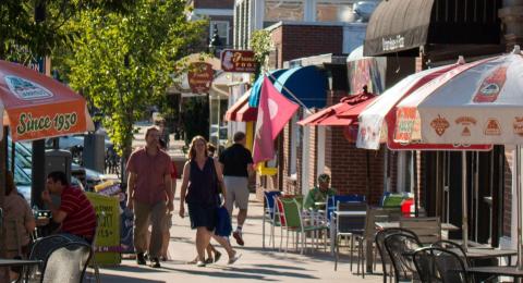 a busy sidewalk with storefronts, outdoor seating, tables and umbrellas