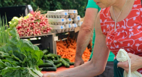 a woman wearing a sleeveless red blouse reaching for fresh greens at a farmers market