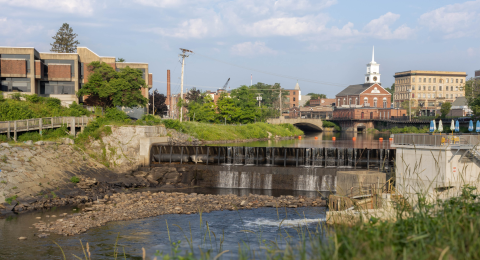 A small urban river with a low dam and waterfall flowing through a town with buildings and a church steeple in the background. The scene includes greenery along the riverbanks, a pedestrian walkway on the left, and a bridge crossing the river near the center.