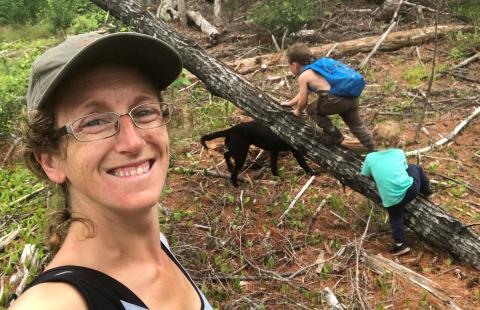 Rachel Dandeneau taking a selfie in the woods with kids and a dog playing behind her