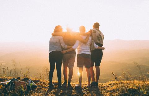 Group of four friends with arms wrapped around their backs looking out across the field at sunrise.