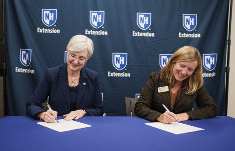 Two women signing papers at a table with blue UNH Extension logo banner in the background