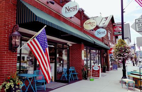 a brick storefront with a green awning, and multiple signs. The scene includes an American flag, blue chairs and tables outside, a hanging flower basket, and a street sign prohibiting skateboards and inline skates on sidewalk.