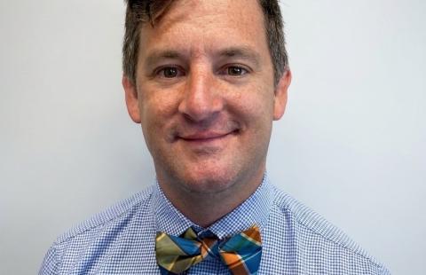 A man wearing a blue checkered shirt and a colorful plaid bow tie, smiling against a plain light gray background. 