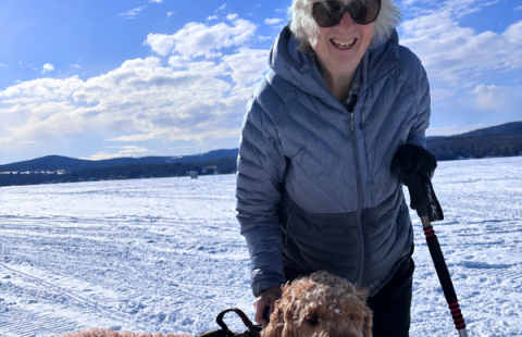 A woman standing on snow-covered ground holding a ski pole and petting a curly-haired dog wearing a harness. Bright blue sky with scattered clouds and distant hills provide a scenic outdoor winter setting.