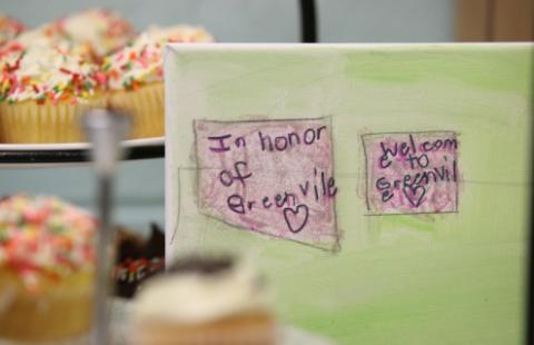 A picture colored by a child that says "In Honor of Greenville" and "Welcome to Greenvile." The words are in purple crayon in a purple colored box; the background is green. Behind the paper is a tray of cupcakes.