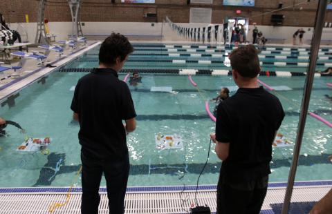 Two youth participate in the Seacoast SeaPerch Challenge. They are both in black shirts, their backs to the camera, looking into an indoor pool at their underwater remote-controlled vehicle.