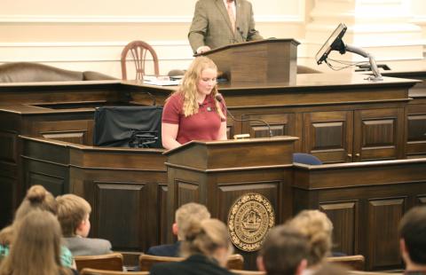 A young woman with blonde hair and wearing a maroon shirt stands at a podium in the NH Statehouse. She is speaking into a microphone. Audience members can be seen in the foreground of the photo.