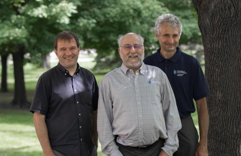 Three men, one in a blue collard shirt on the left, one in a light blue shirt and with a beard and glasses and the center, and one on the right in a blue polo shirt and standing next to a tree, smile and face the camera.
