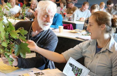 Two people are looking at a plant. They are seated with others in a classroom. On the right, a woman in a gray shirt looks at and identifies the plant by holding a leaf. On the left, a man with a white beard and a blue shirt holds the plant and is looking at the woman and listening to her.