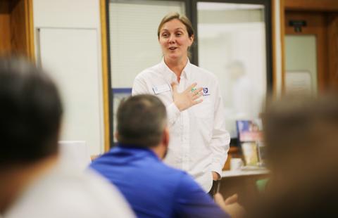 A woman in a white shirt addresses an audience in a classroom. The audience is visible in the foreground of the photo. She is smiling and holding her hand to her chest.