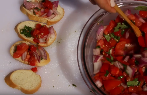 Bowl of freshly made tomato bruschetta, being spooned onto small rounds of bread.
