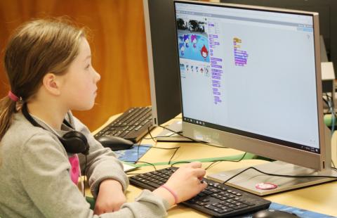 A young girl is looking at a computer; on the screen is some computer code and some animated characters.
