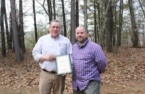 Two men stand next to each other in the woods. Behind them are trees. The man on the left is wearing a white shirt and holding a plaque. The man on the right is wearing a purple plaid shirt. They are both looking at the camera.