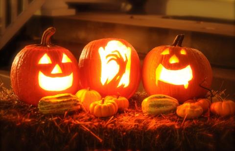Three jack-o-lanterns on a bale of hay. The jack-o-lanterns have faces carved into them and they are lit by candles. Smaller decorative gourds surround them.