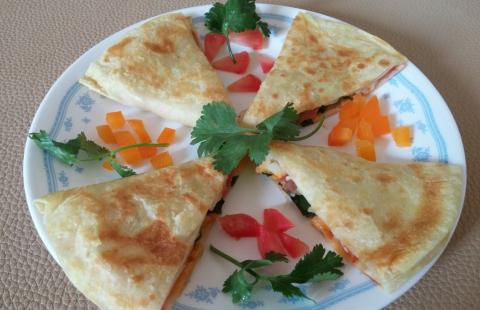 A quesadilla cut into four slices and arranged in a cross shape on a plate. Chopped orange and red peppers and pieces of parsley are between the slices. The plate is white with a blue pattern.