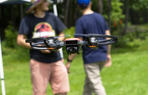 A close-up photo of a drone hovering in the air. It has a pair of rotary blades on each side. In the lower center of the drone is a camera. Behind the drone stand two men. One is holding the controls for the drone.