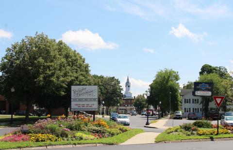 A street scene in downtown Rochester NH. In the foreground is a traffic island that is filled with a flower garden and a sign for the Rochester Opera House. In the center, there is a street with cars on it. On either side of the street are trees and buildings. In the distance is a white steeple.