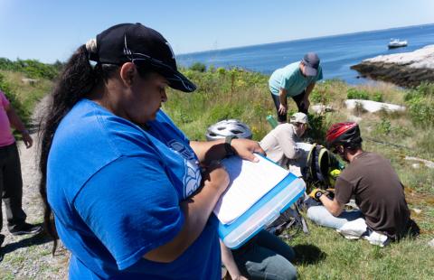A woman in a blue shirt and cap is writin gsomething on a clipboard. Behind her, five volunteers are engaged in activities on a rocky, grassy hillside.