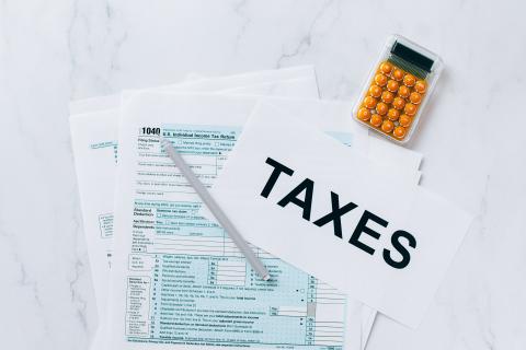 Stock image of tax forms with the word "taxes" in large black print. An orange calculator sits next to the forms.