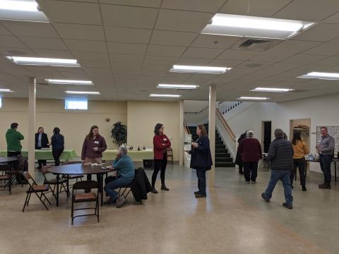 Several event participants mingle in a large room. At center, there is a round table with people sitting and talking