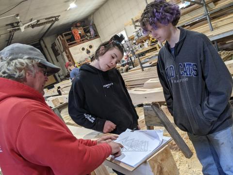 Instructor and two interns discussing architectural drawings in a woodworking shop.