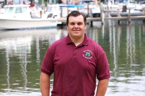 Teen boy wearing 4-H shirt, standing in front of water with boats in background