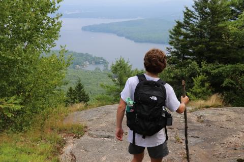 A young person with a black backpack and walking stick stands atop a mountain over looking lake Winnipesaukee