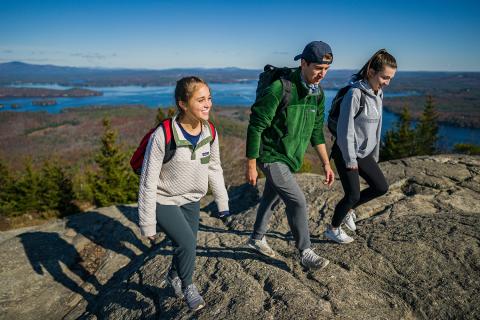 3 Young people with backpacks hike up a mountain with a lake in the distance