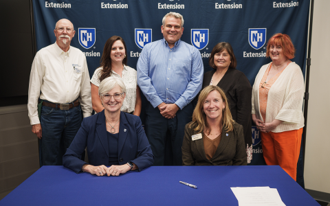 Group of people posing in front of a blue banner with UNH Extension logos for an official signing. Two women are seated at the table in the foreground.