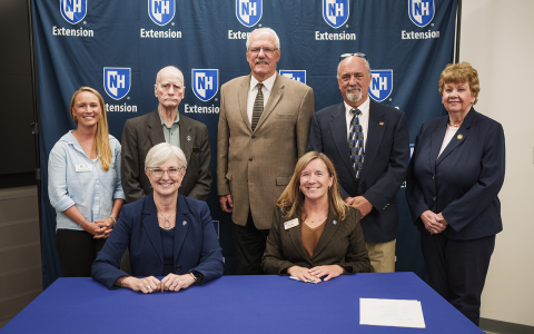 Group of people posing in front of a blue banner with UNH Extension logos for an official signing. Two women are seated at the table in the foreground.