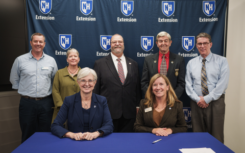 Group of people posing in front of a blue banner with UNH Extension logos for an official signing. Two women are seated at the table in the foreground.