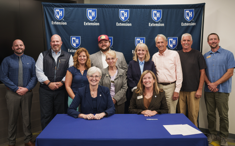 Group of people posing in front of a blue banner with UNH Extension logos for an official signing. Two women are seated at the table in the foreground.