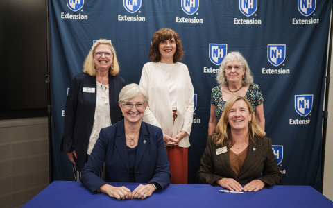 Group of people posing in front of a blue banner with UNH Extension logos for an official signing. Two women are seated at the table in the foreground.