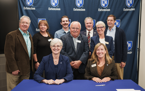Group of people posing in front of a blue banner with UNH Extension logos for an official signing. Two women are seated at the table in the foreground.
