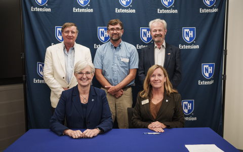 Group of people posing in front of a blue banner with UNH Extension logos for an official signing. Two women are seated at the table in the foreground.