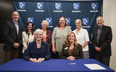 Group of people posing in front of a blue banner with UNH Extension logos for an official signing. Two women are seated at the table in the foreground.