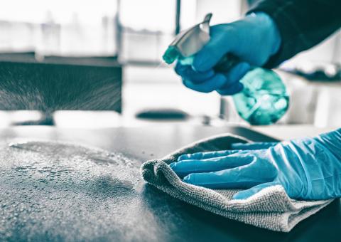 gloved hands, one hand spraying cleaner onto food preparation surface, the other holding microfiber cloth