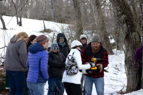 a group of women look on while a women wearing a white coat learns how to drill a hole into a sugar maple to collect sap