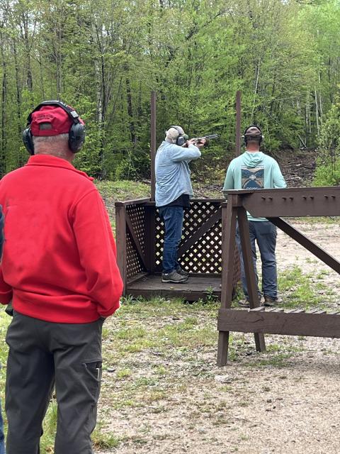 an adult shoots a shotgun, two others look on from behind